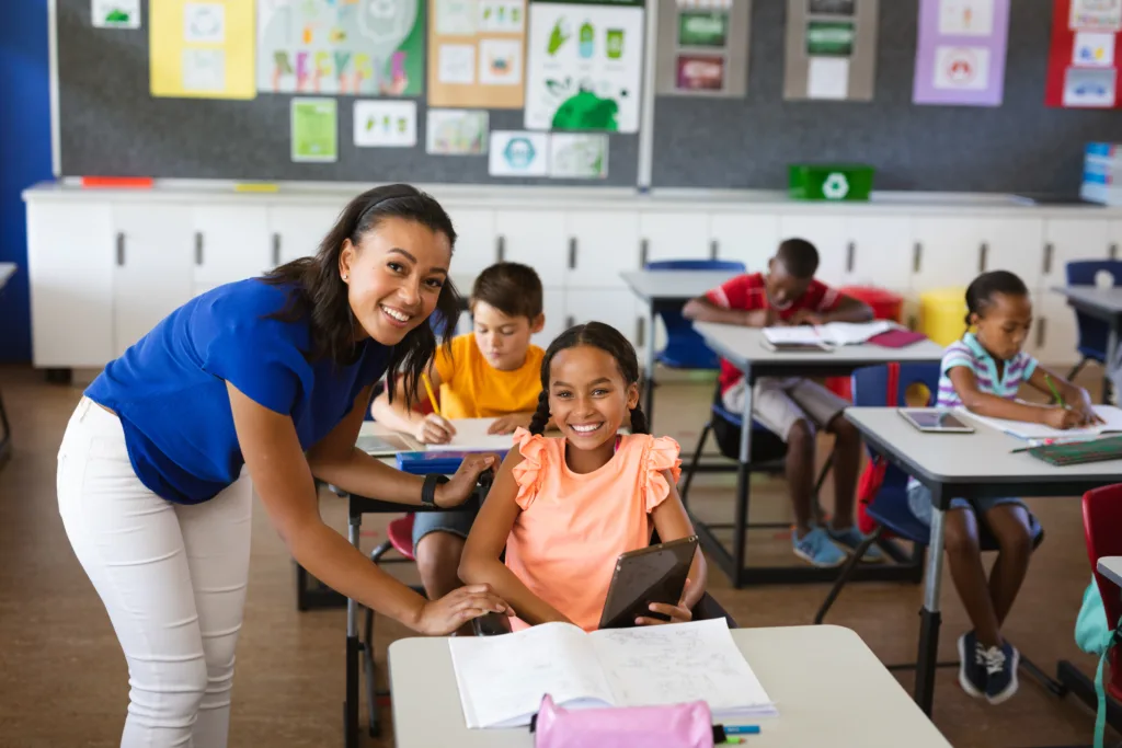 Young student at her desk in classroom with paraprofessional helping her