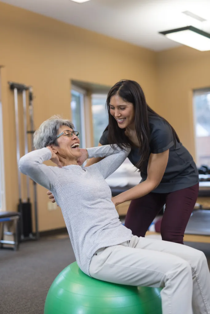 A female occupational therapist works with a cheerful senior female as she does repetitions on an exercise ball.