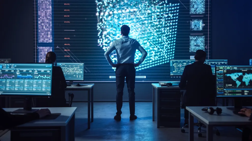 Project manager Stands Next to Big Screen while Team of Professional Computer Data Science Engineers Work on Desktops in a Dark System Control Office.