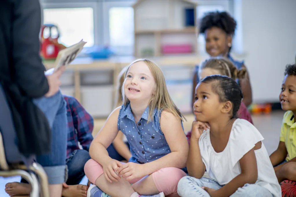 elementary school children are seated together on a classroom floor listening intently as their teacher reads them a story from a book