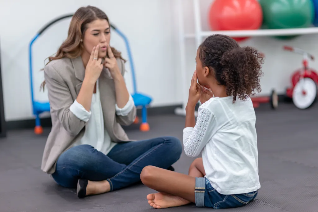 A pediatric occupational therapist is doing rehab work with a young girl in her clinic. The girl is sitting across from the therapist and mirroring what she's doing.