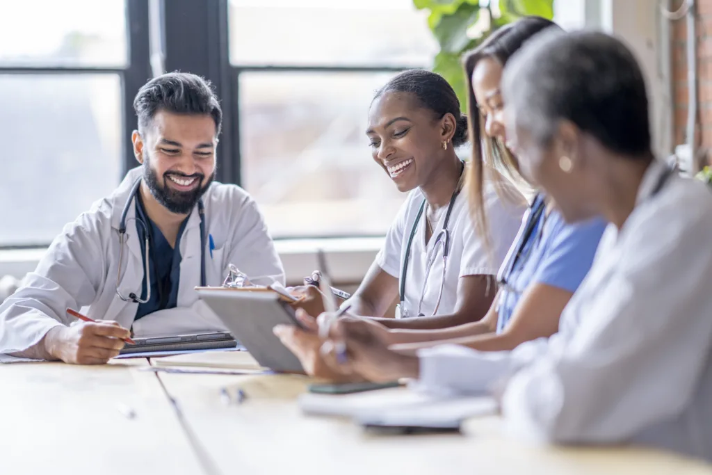 A small group of four medical professionals sit around a boardroom table as they meet to discuss patient cases.  They are each dressed professionally in scrubs and lab coats as they focus on working together.