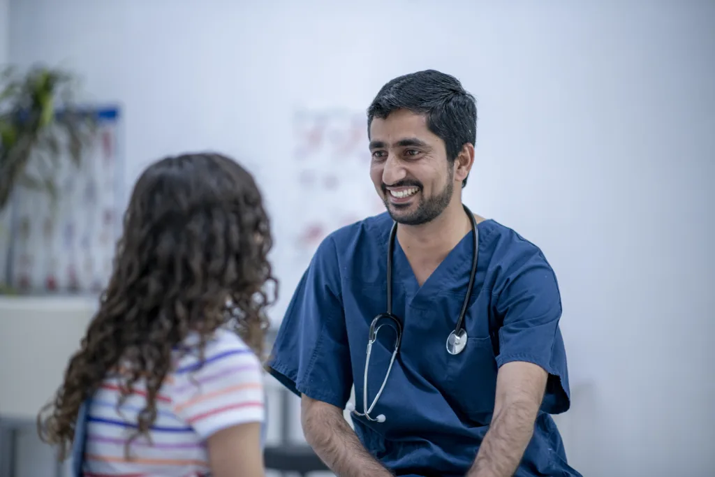 A male doctor sits in an exam room with a young female patient as they talk about her health