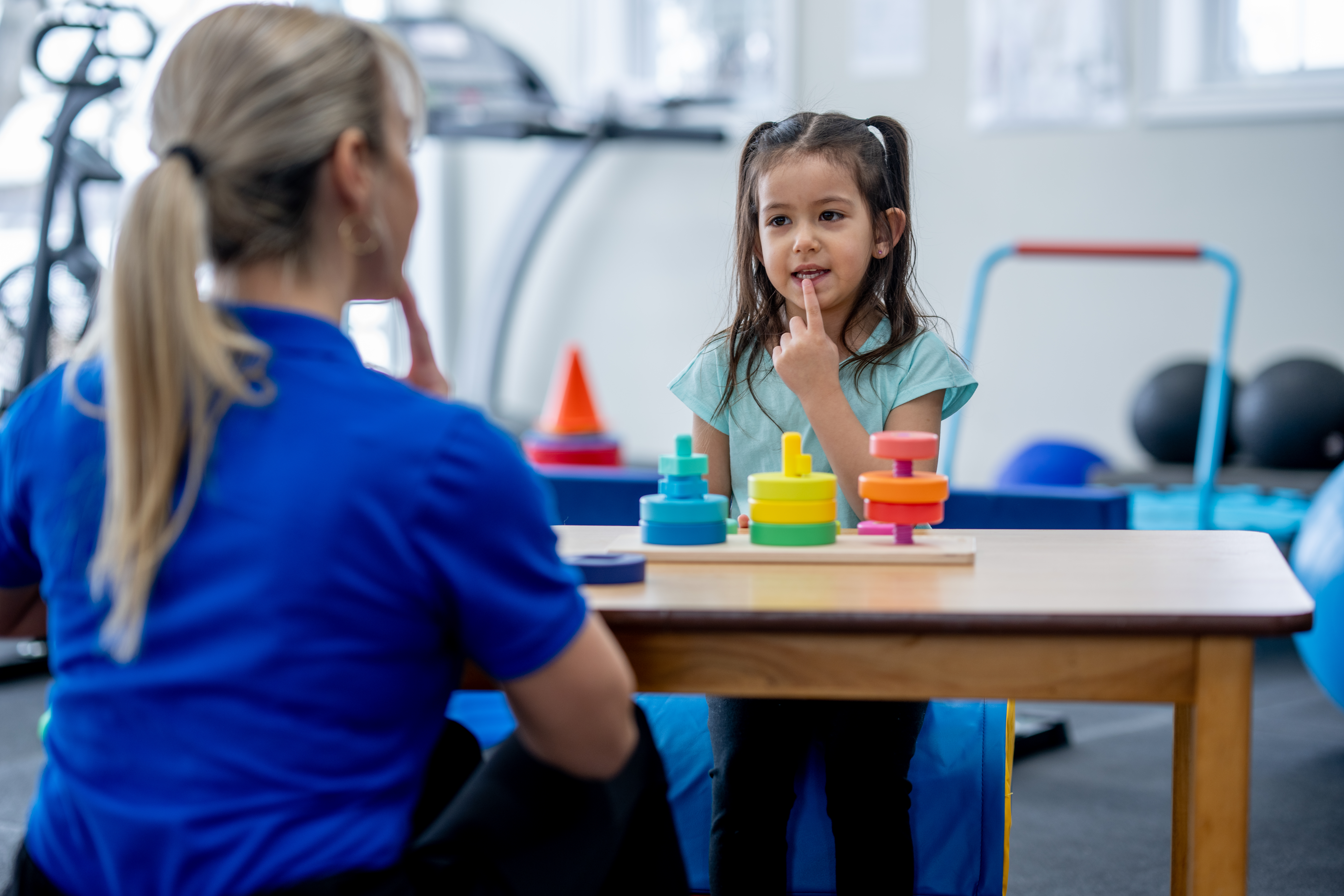 A female Speech Therapist works with a little girl on forming her letters and sounds.  The two are seated at a small wooden table facing each other as the little girl mimics the therapists sounds.
