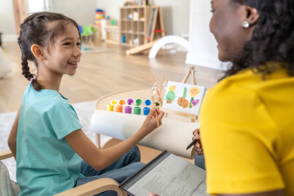 A female therapist sits at a table with a little girl during an art therapy session.  