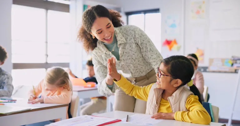 Mujer profesora, niña choca los cinco y aula con logros, éxito y tutoría para el aprendizaje. La educación, el desarrollo y los estudiantes con metas, conocimientos y libros con la celebración en el escritorio de la escuela.
