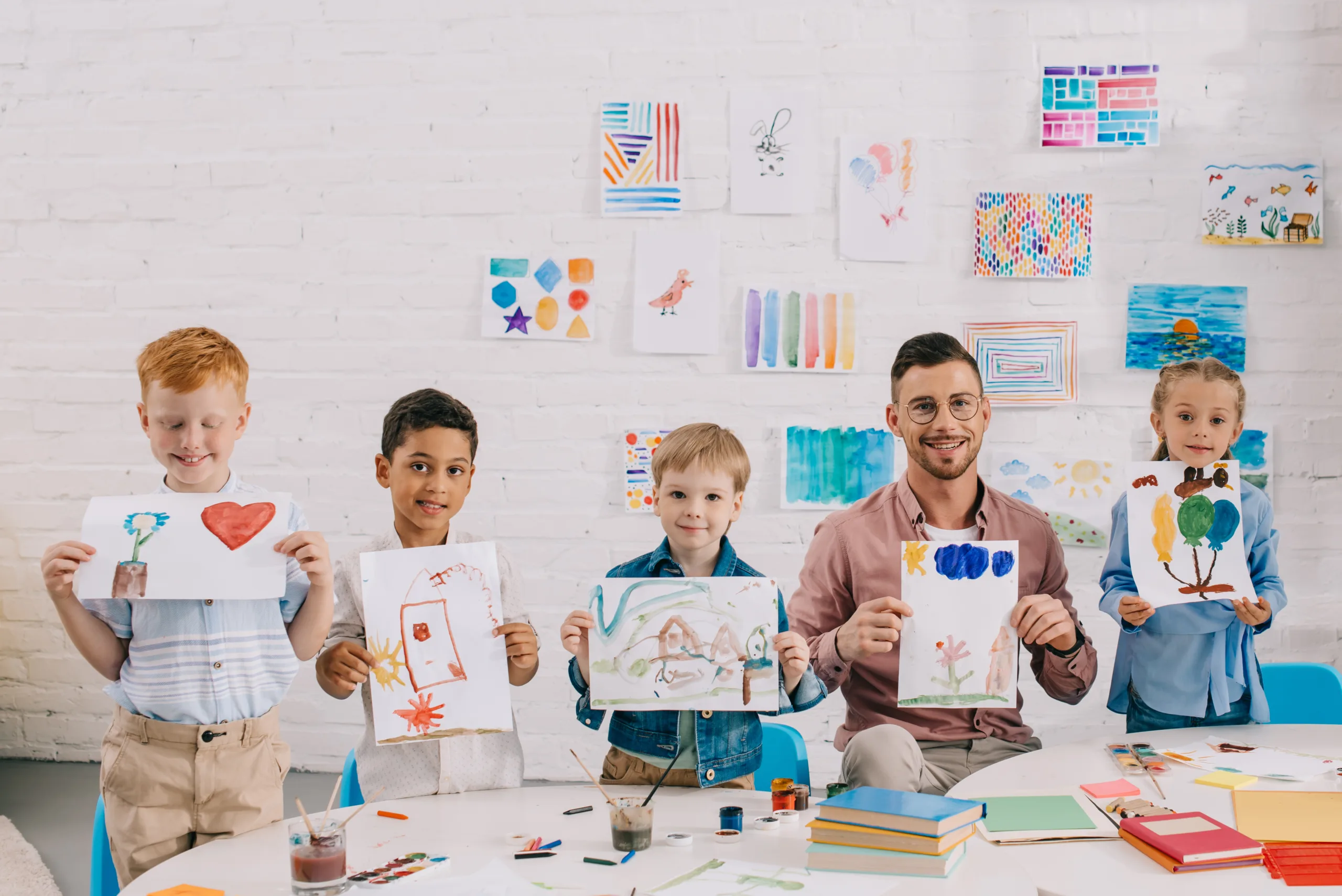 portrait of smiling teacher and multicultural preschoolers showing colorful pictures in hands in classroom