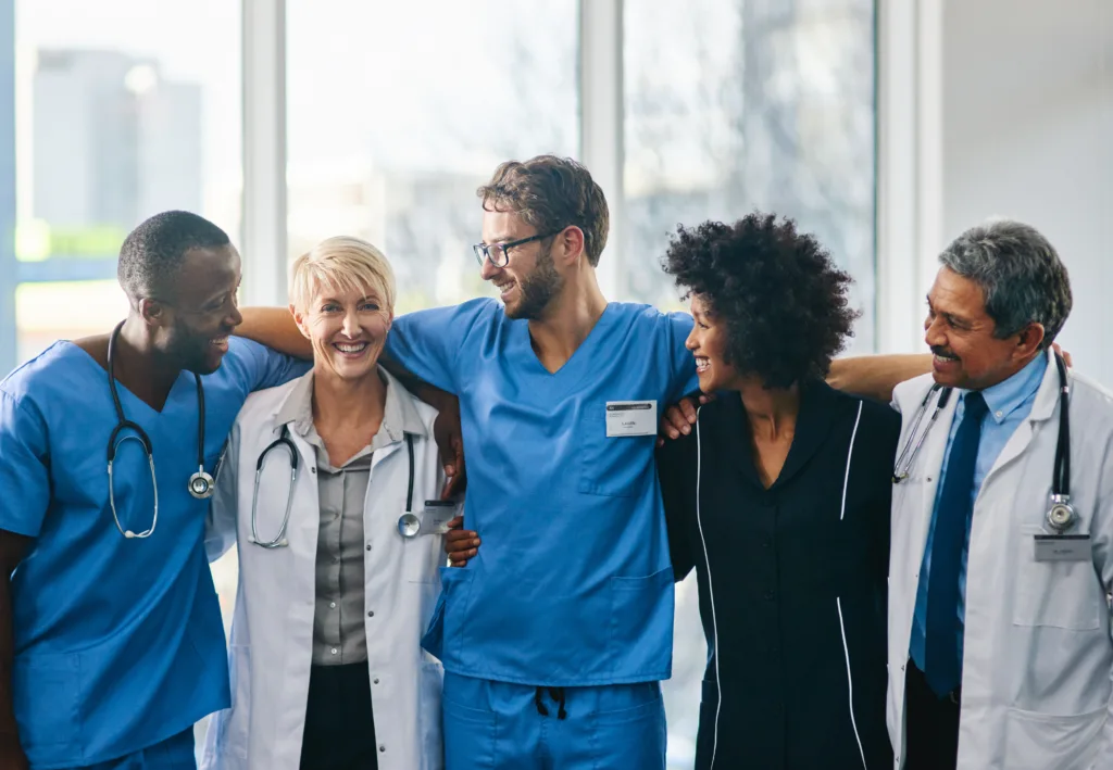 A diverse group of smiling healthcare professionals standing together, showing unity and support. They are dressed in scrubs and lab coats, representing teamwork, wellness, and positivity in the medical field.