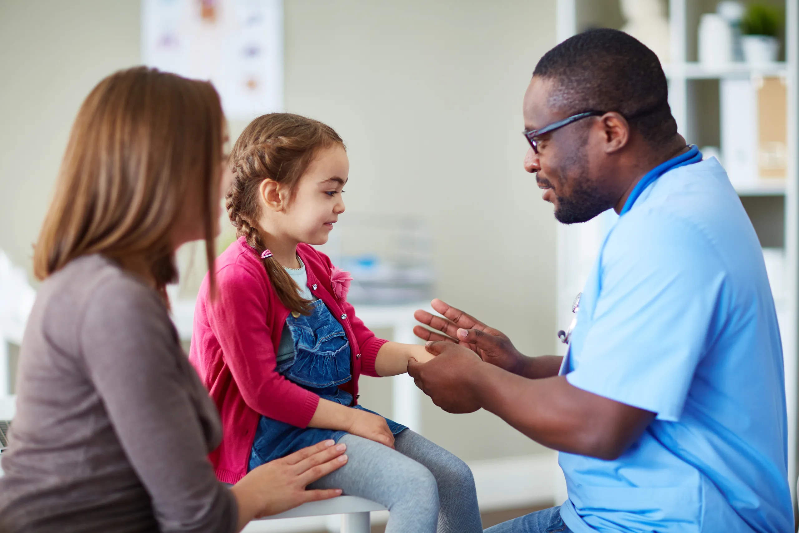 Eine Krankenschwester untersucht die Hand eines kleinen Mädchens, während ihre Mutter in einer Kinderklinik neben ihr sitzt.